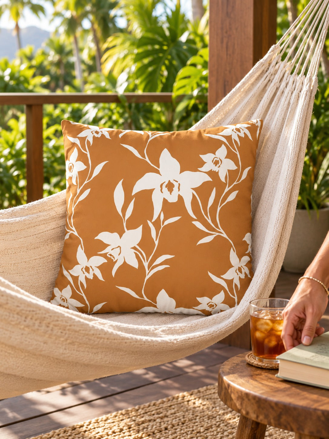 A square decorative throw pillow with an orchid floral vine pattern on a warm rust-orange background, styled inside a cream-colored hammock on a wooden deck with tropical plants in the background.