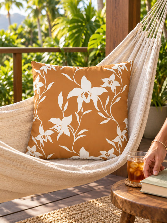 A square decorative throw pillow with an orchid floral vine pattern on a warm rust-orange background, styled inside a cream-colored hammock on a wooden deck with tropical plants in the background.
