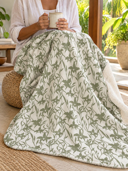 Woman enjoying morning coffee on a tropical lanai with an Orchid Sage sherpa fleece blanket draped across her lap.