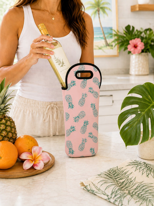 A woman in a white tank top holds a wine bottle next to a pink insulated wine bag with a blue pineapple pattern, set on a kitchen counter with tropical fruits and plants.
