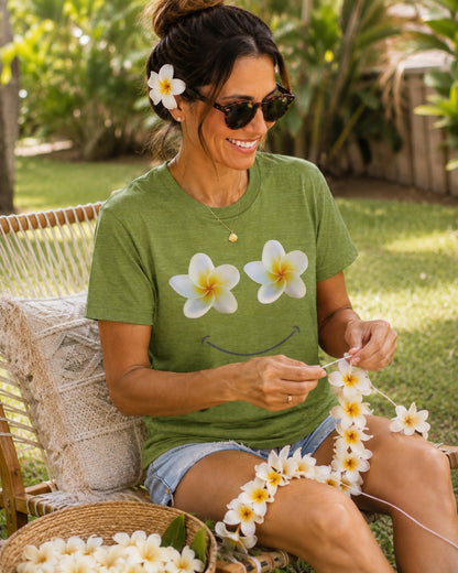 A woman sitting outdoors on a woven chair, wearing an olive green t-shirt with a white and yellow plumeria flower smiley face graphic. She is holding a white flower lei and has plumeria flowers in a basket in the foreground.