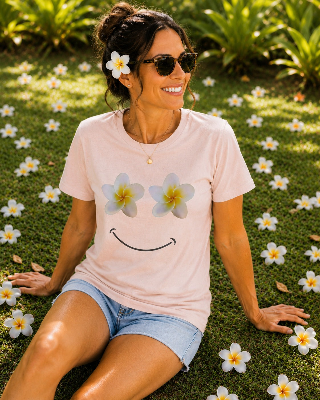 A woman sitting on green grass outdoors, wearing a light pink short-sleeve T-shirt with a smiley face made from plumeria flowers for eyes and a curved smile. She is also wearing denim shorts, sunglasses, and a flower in her hair.