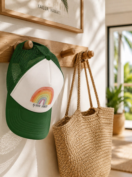 A green and white trucker hat with a rainbow graphic and the text 'ALOHA VIBES' hangs on a wooden wall rack next to a woven straw tote bag in a bright, sunlit room.