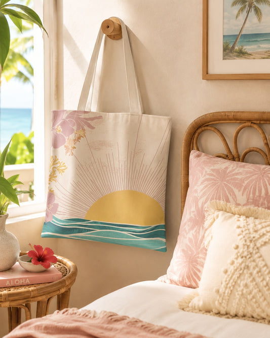 A canvas tote bag with a tropical sunset and floral print hangs on a wooden wall hook in a sunlit bedroom. The bag features a cream base with a yellow sun, teal ocean waves, and pink flowers.