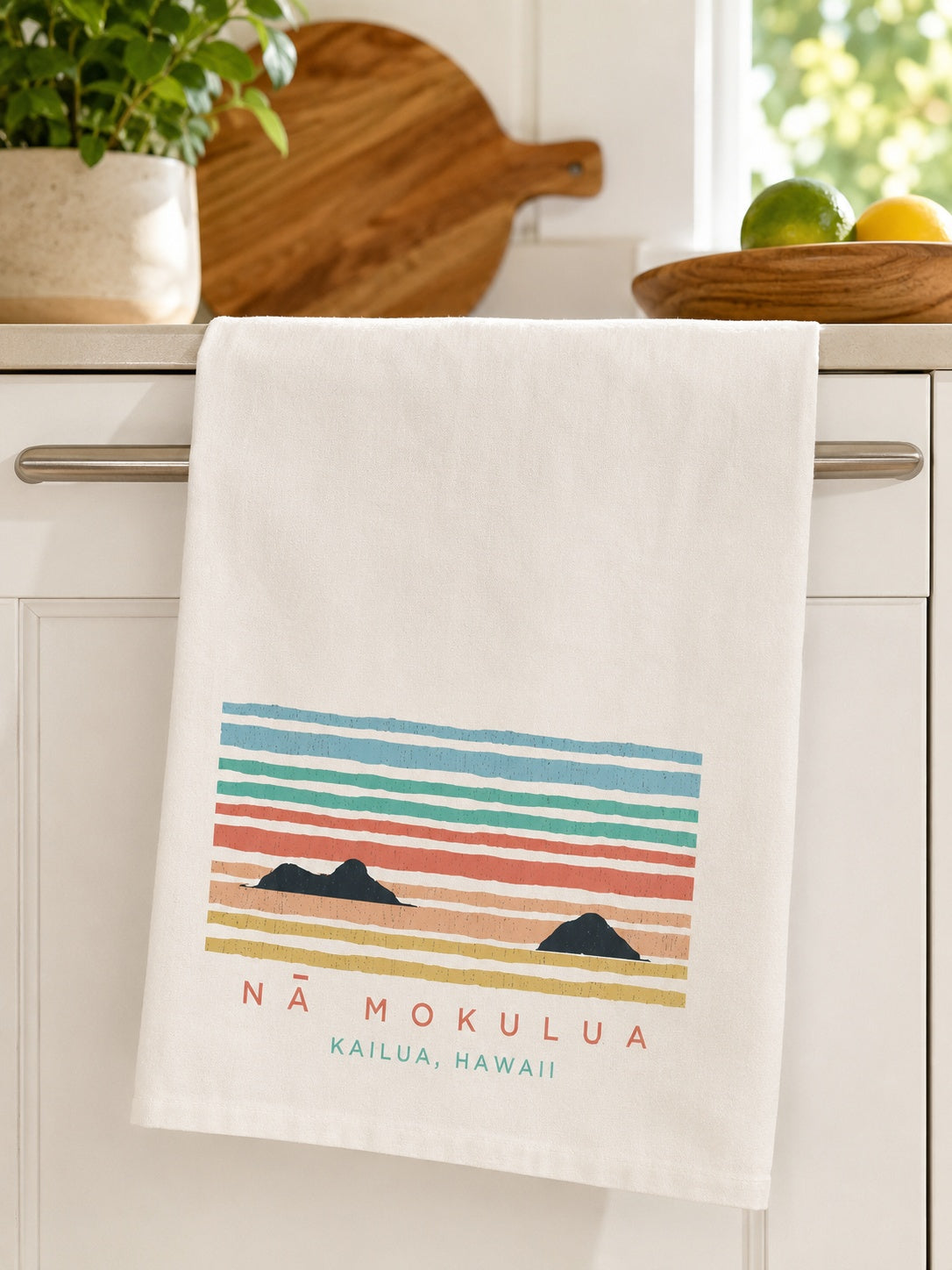 A white kitchen towel hanging from a silver cabinet handle, featuring a colorful horizontal striped sunset-style graphic with two black mountain silhouettes and the text 'NÁ MOKULIA KAILUA, HAWAII'. A kitchen counter with a cutting board, plant, and fruit is visible in the background.