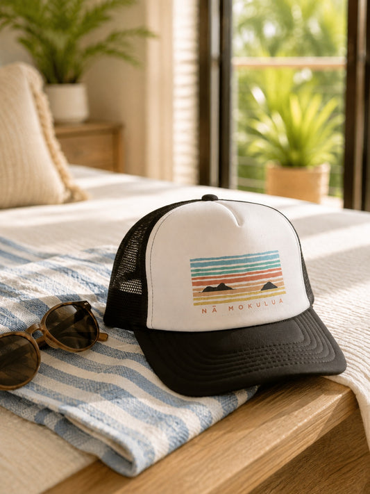 A black and white trucker hat with a colorful sunset graphic and 'NA MOKULUA' text, resting on a blue and white striped towel on a wooden bed frame in a sunlit room.